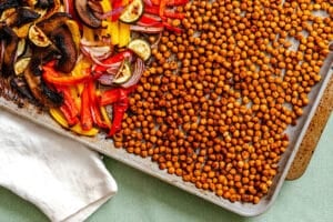 A baking sheet with roasted chickpeas on one side and roasted vegetables including red onions, bell peppers, zucchini, and mushrooms on the other side.