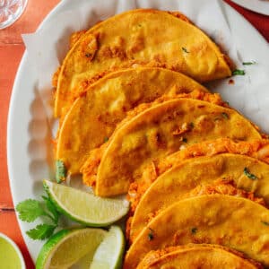 A plate of crispy tacos filled with a reddish-orange filling, garnished with cilantro, served with lime wedges and green sauce on the side.