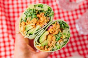 A hand holds a wrap cut in half, showing lettuce, cucumber, onions, avocado, and a creamy filling, against a red and white checkered background.