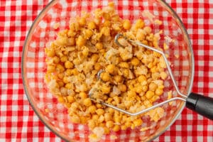 A glass bowl of chickpeas is being mashed with a metal potato masher on a red and white checkered tablecloth.