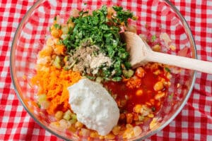 A glass bowl with chickpeas, grated carrot, chopped herbs, white sauce, red sauce, and spices being mixed with a wooden spoon on a red checkered tablecloth.