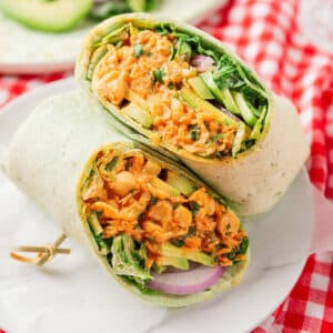 A wrap sandwich sliced in half, filled with shredded vegetables, greens, and a creamy orange mixture, placed on a white plate over a red and white checkered cloth.