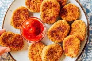 A plate of breaded nuggets arranged around a small glass bowl of ketchup, with one nugget being picked up by hand.