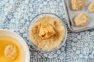 A bowl of bread crumbs with a ball of dough on top, next to a bowl of liquid and a tray with breaded nuggets on a patterned cloth.