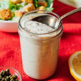 A glass jar of creamy Tahini Caesar Dressing with a spoon inside, set on a red cloth, with a salad, lemon slice, and capers in the background.