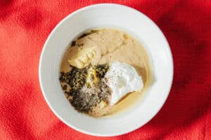 A white bowl contains Tahini Caesar Dressing made with tahini, yogurt, mustard, chopped herbs, and ground black pepper, placed on a red textured surface.