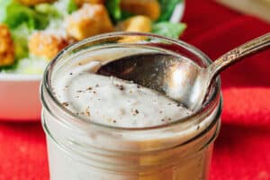 A close-up of a jar of creamy Tahini Caesar Dressing with black pepper on top, and a spoon inside. A bowl of salad with croutons is blurred in the background.