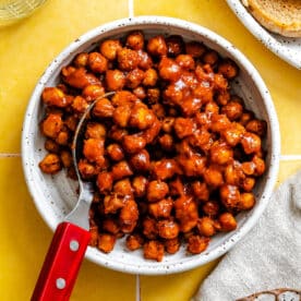 A white bowl filled with barbecue-coated chickpeas sits on a yellow tile surface, with a metal spoon featuring a red handle resting inside the bowl.