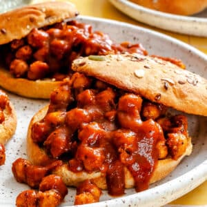 Two open sandwiches with chickpea and tomato-based sauce filling on seeded buns, served on a speckled plate.