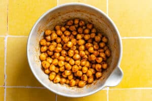 A white bowl filled with seasoned chickpeas sits on a yellow tiled surface.