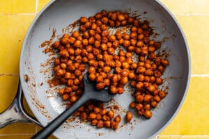 Chickpeas coated in spices being stirred in a gray skillet with a black spoon on a yellow tiled surface.