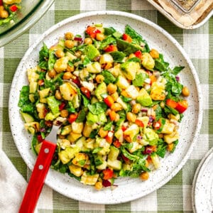 A plate of vibrant halloumi chickpea salad with spinach, avocado, corn, red pepper, and cucumber sits on a checkered tablecloth, accompanied by a fork and glass.