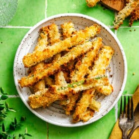 A plate of crispy, breaded zucchini fries on a white dish, with more fries on a wooden board nearby, set on a green tiled surface. Fork and yellow napkin are visible.
