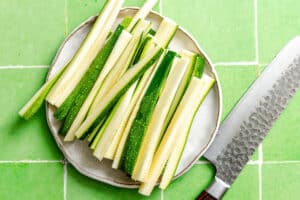 A plate of raw zucchini sticks next to a large knife on a green tiled surface.