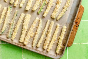 Breaded zucchini sticks arranged in rows on a parchment-lined baking sheet, ready to be baked, with the tray placed on a green tiled surface.