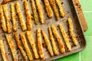 A baking tray with rows of golden-brown, breaded zucchini fries arranged on parchment paper.