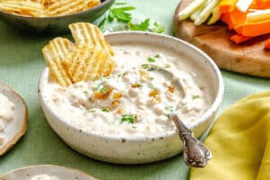 A bowl of creamy onion dip garnished with herbs, with a spoon and two potato chips, next to a platter of sliced vegetables.
