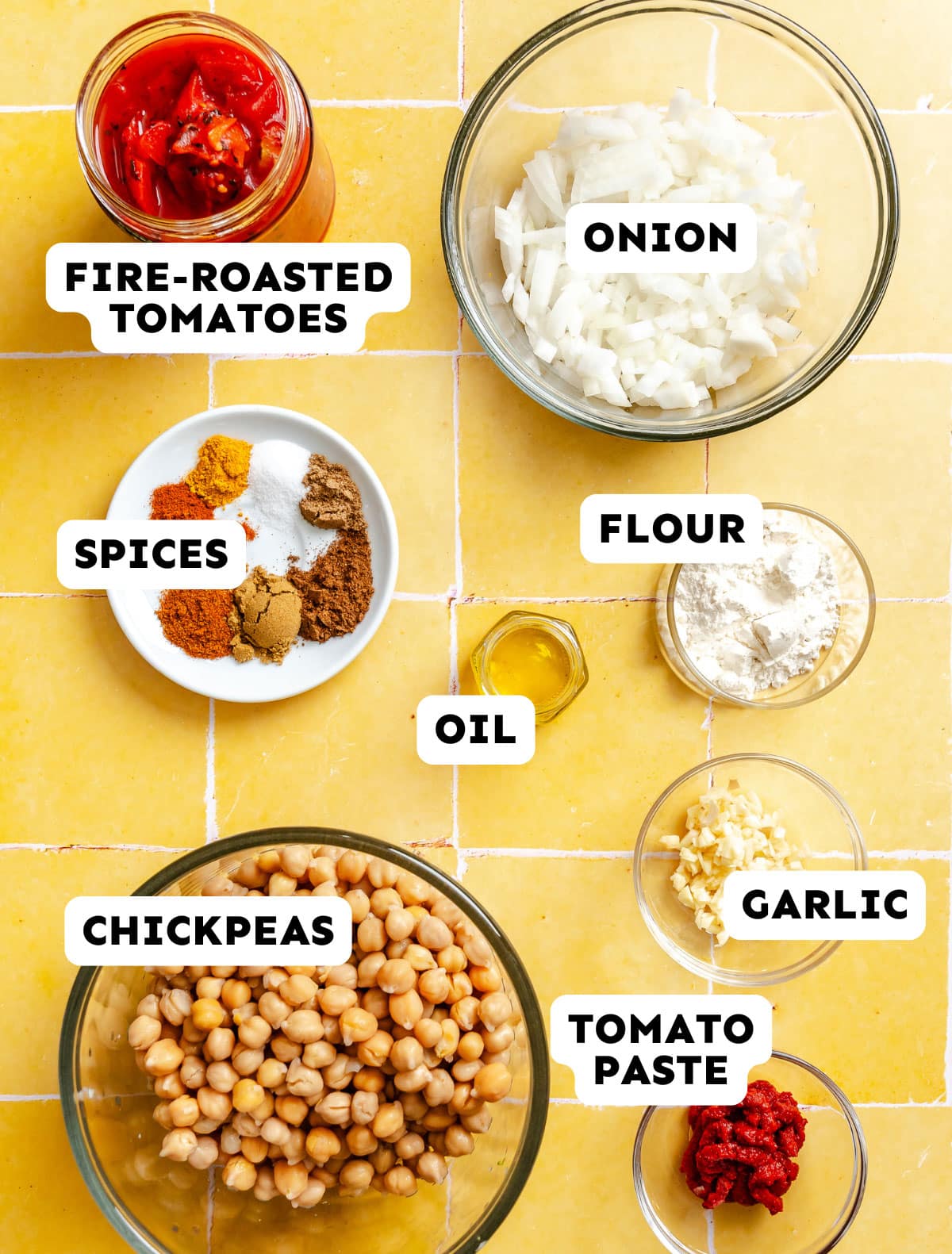Bowls containing fire-roasted tomatoes, Moroccan chickpeas, chopped onion, spices, flour, oil, minced garlic, and tomato paste are arranged on a yellow tiled surface.