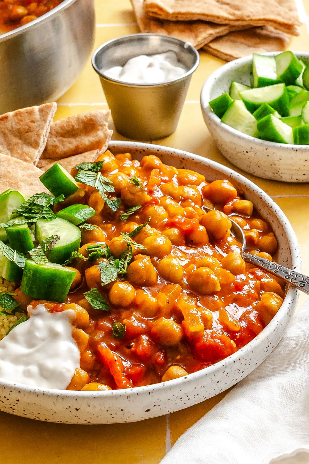 A bowl of chickpea stew garnished with cucumber, mint, and a dollop of yogurt, with pita bread and extra yogurt and cucumber on the side.