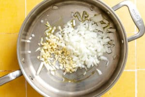 Chopped onions and garlic in a stainless steel pan with some oil, on a yellow tiled countertop.