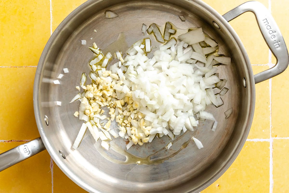 Chopped onions and garlic in a stainless steel pan with some oil, on a yellow tiled countertop.