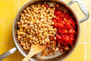 A stainless steel pan containing cooked onions, garlic, chickpeas, and chopped tomatoes, with a wooden spoon resting inside on a yellow tiled surface.