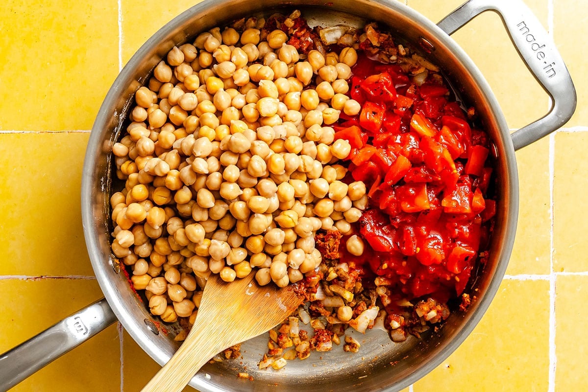 A stainless steel pan containing cooked onions, garlic, chickpeas, and chopped tomatoes, with a wooden spoon resting inside on a yellow tiled surface.