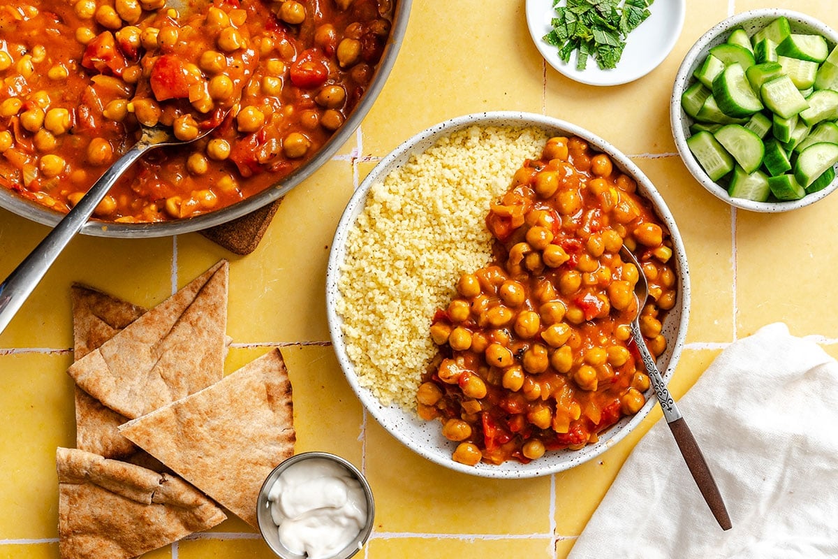 A bowl of couscous topped with chickpea stew sits beside pita bread, cucumber slices, fresh herbs, and a small dish of sauce on a yellow tiled surface.