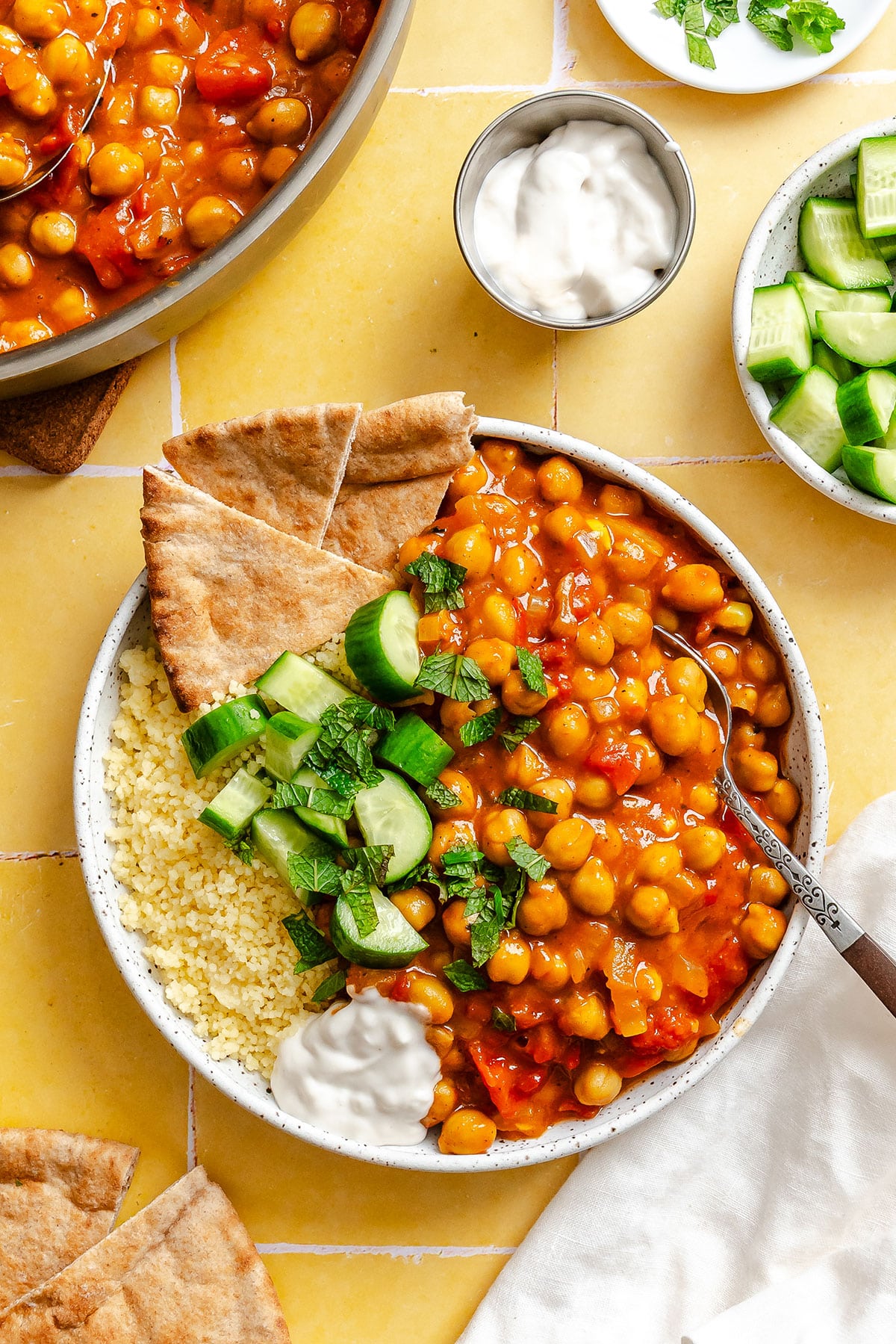 A bowl with chickpea stew, couscous, cucumber slices, pita bread, and a dollop of white sauce, photographed overhead on a yellow tiled surface.