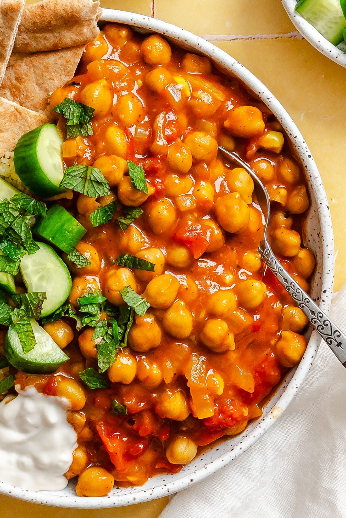 A bowl of chickpea stew with tomato sauce, garnished with fresh mint, sliced cucumber, pita bread, and a dollop of yogurt.