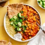 A bowl contains chickpea stew, couscous, pita bread, cucumber pieces with mint, and a dollop of white sauce on a yellow tile surface.