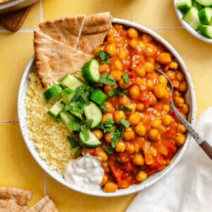 A bowl contains chickpea stew, couscous, pita bread, cucumber pieces with mint, and a dollop of white sauce on a yellow tile surface.