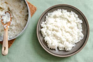A plate of cooked white rice sits next to a pot with more rice and a wooden spoon, all on a light green tablecloth.