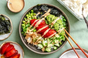 A bowl of rice topped with sliced avocado, marinated tomatoes, edamame, cucumbers, green onions, nori, and sesame dressing, with chopsticks placed on the rim.