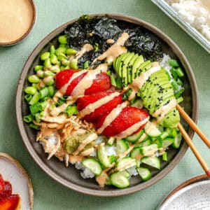 A bowl of rice topped with sliced avocado, marinated tomatoes, edamame, cucumbers, green onions, nori, and sesame dressing, with chopsticks placed on the rim.