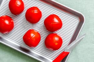 Six whole tomatoes with X-shaped cuts on top are arranged on a baking sheet next to a paring knife with a red handle, on a light green surface.