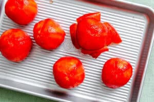 Six peeled tomatoes, one partially peeled, are arranged on a ridged metal baking sheet.