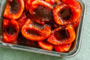A glass dish with halved red tomatoes covered in a brown liquid marinade, placed on a green textured surface.