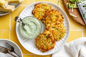 Three corn fritters are served on a white plate with a small jar of creamy green dipping sauce; fresh corn and grated zucchini are visible nearby.