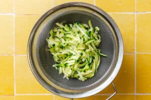 Shredded zucchini sits in a metal strainer placed over a yellow tiled surface.