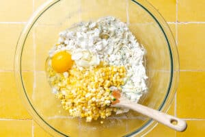 A clear glass bowl contains flour, a cracked egg, corn kernels, chopped green onions, and crumbled cheese, with a wooden spoon resting inside.
