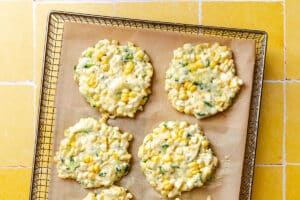 Five uncooked zucchini corn fritters sit on a parchment-lined baking rack over a yellow tiled surface.