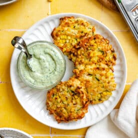 Three zucchini fritters are served on a white plate with a small glass bowl of creamy green dipping sauce and a spoon, placed on a yellow tiled surface.