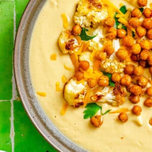 A bowl of creamy cauliflower soup topped with roasted cauliflower, crispy chickpeas, shredded cheese, and parsley, set on a green tiled surface.