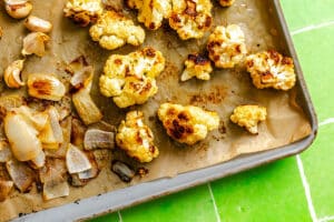 Roasted cauliflower florets and chopped onions on a parchment-lined baking sheet, resting on a bright green tiled surface.