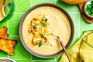 A bowl of creamy cauliflower soup topped with roasted cauliflower, shredded cheese, and herbs, with a spoon inside. Toast, bread, and a garnish of chopped parsley are nearby on a green tiled surface.