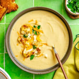 A bowl of creamy cauliflower soup garnished with roasted cauliflower, shredded cheese, and parsley sits on a green tiled surface with toast, herbs, and bread nearby.