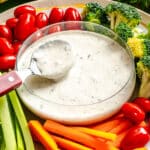 A bowl of creamy ranch dip with a serving spoon, surrounded by fresh vegetables including broccoli, cherry tomatoes, celery, and carrot sticks.