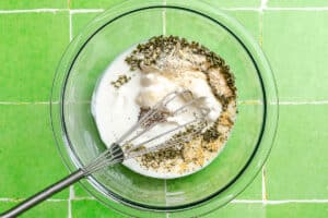 A glass bowl on a green tiled surface contains milk, mayonnaise, herbs, and seasoning with a metal whisk placed inside.
