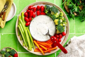 A plate of fresh celery, carrots, broccoli, grape tomatoes, and ranch dip with a spoon on a green tiled surface.
